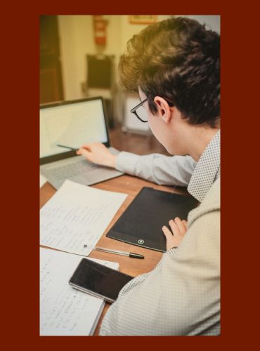 A man with glasses is sitting at a desk and reading from a laptop screen.