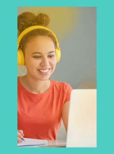A smiling woman wearing headphones and looking at a laptop screen while taking notes on paper.